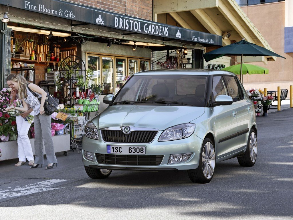 Srebrna Skoda Fabia II zaparkowana w mieście obok dwóch młodych dziewczyn robiących zakupy